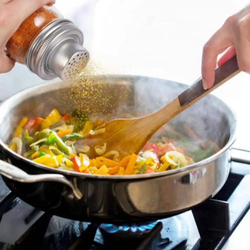 A hand is shaking a stainless steel mason jar lid filled with yellow seasoning over food in a pan.