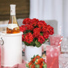 A bouquet of red geranium flowers in a white pot next to a bottle and pink drinks.