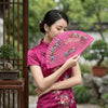 A woman in a pink traditional dress holds and displays a hand-painted decorative folding fan outdoors.