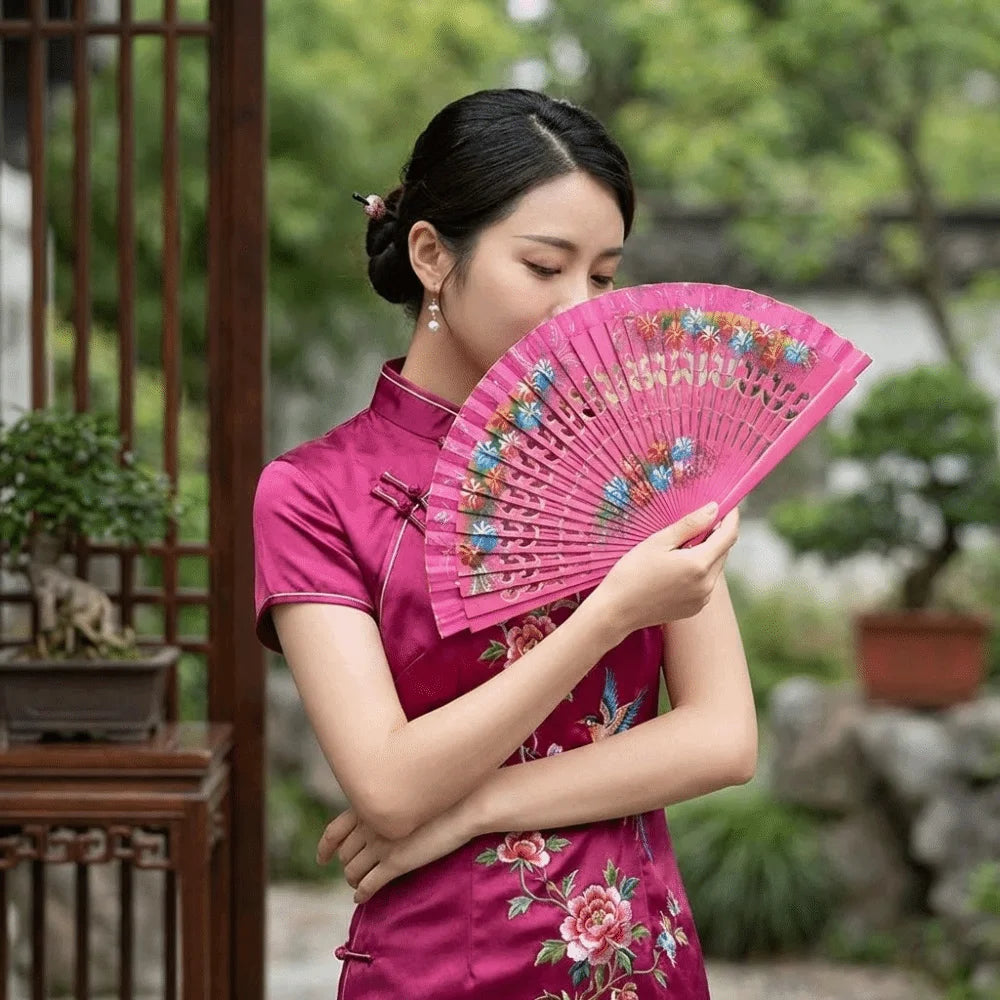 A woman in a pink traditional dress holds and displays a hand-painted decorative folding fan outdoors.