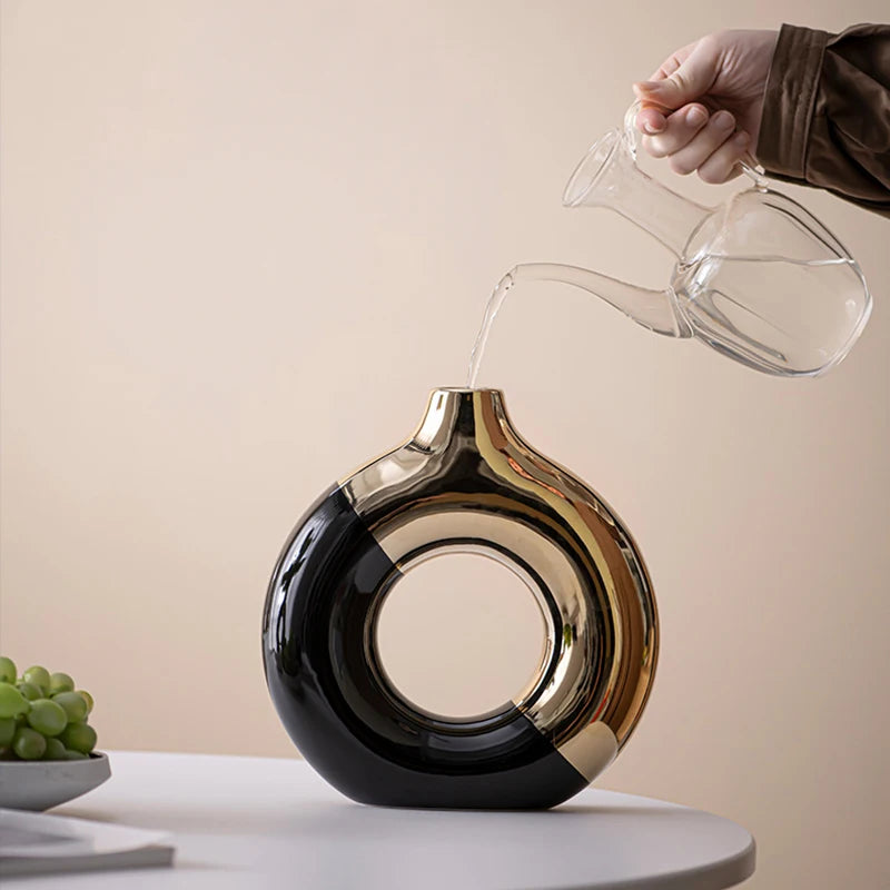 A hand pours water from a clear glass vessel into a black and gold ceramic flower vase.