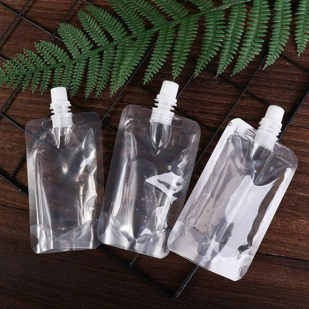 Three clear reusable squeezable liquid drink pouches resting on a dark wooden surface with a green fern leaf in the backgroun