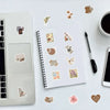 A spiral notebook filled with various incomplete diary style graffiti stickers on a white surface next to a silver laptop.