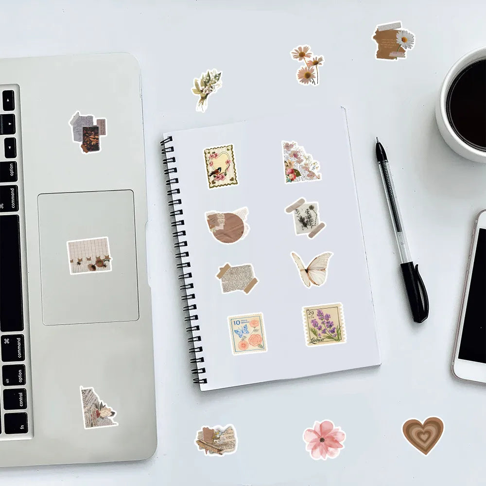A spiral notebook filled with various incomplete diary style graffiti stickers on a white surface next to a silver laptop.