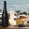 A black champagne bottle stands next to elegant disposable champagne glasses and cheese on a wooden table.