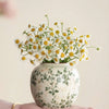 A hand holds a white ceramic vase with green floral patterns filled with small white and yellow daisies.