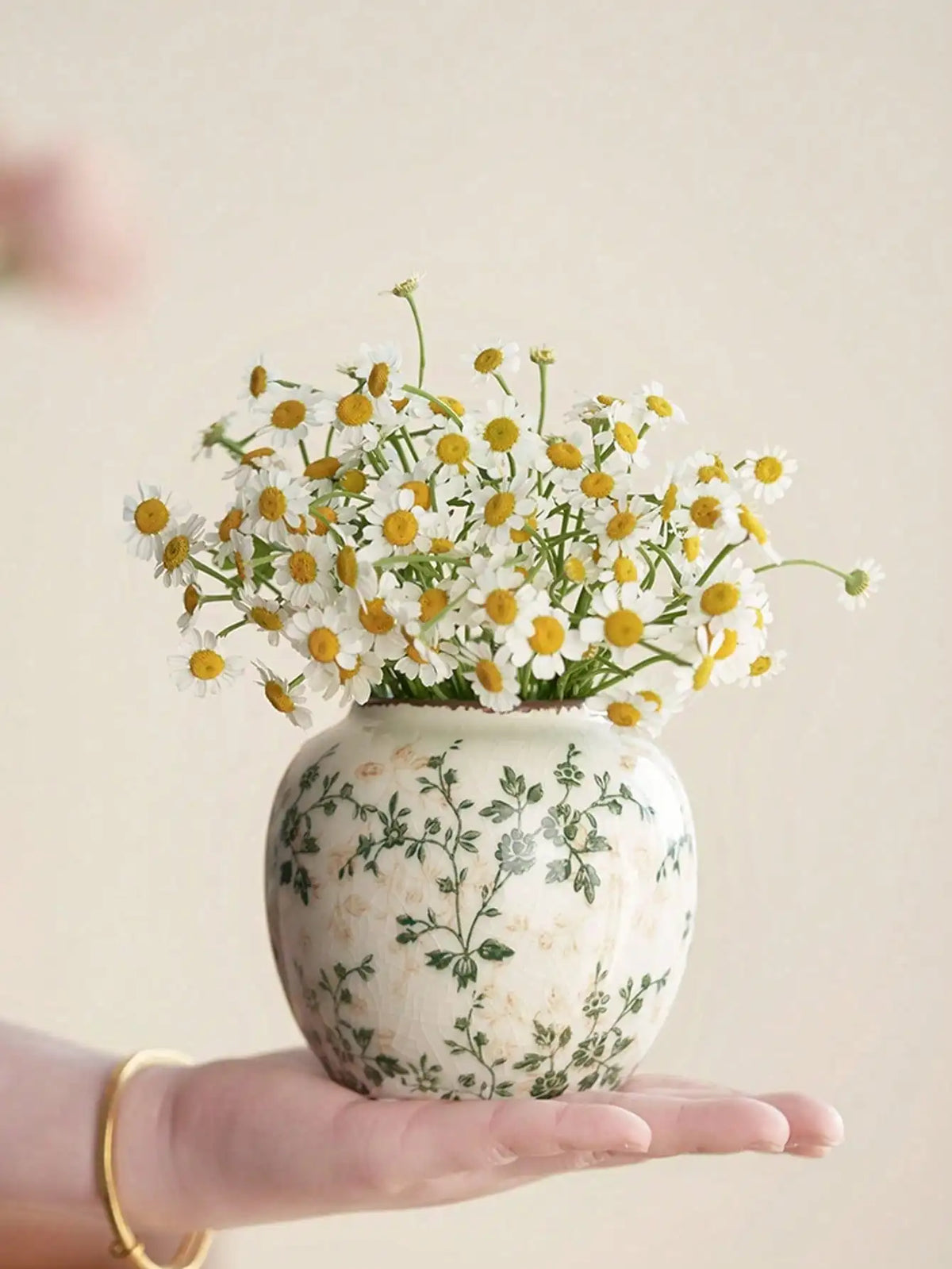A hand holds a white ceramic vase with green floral patterns filled with small white and yellow daisies.