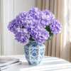 A bouquet of purple artificial hydrangea silk flowers in a blue and white patterned vase on a white table.