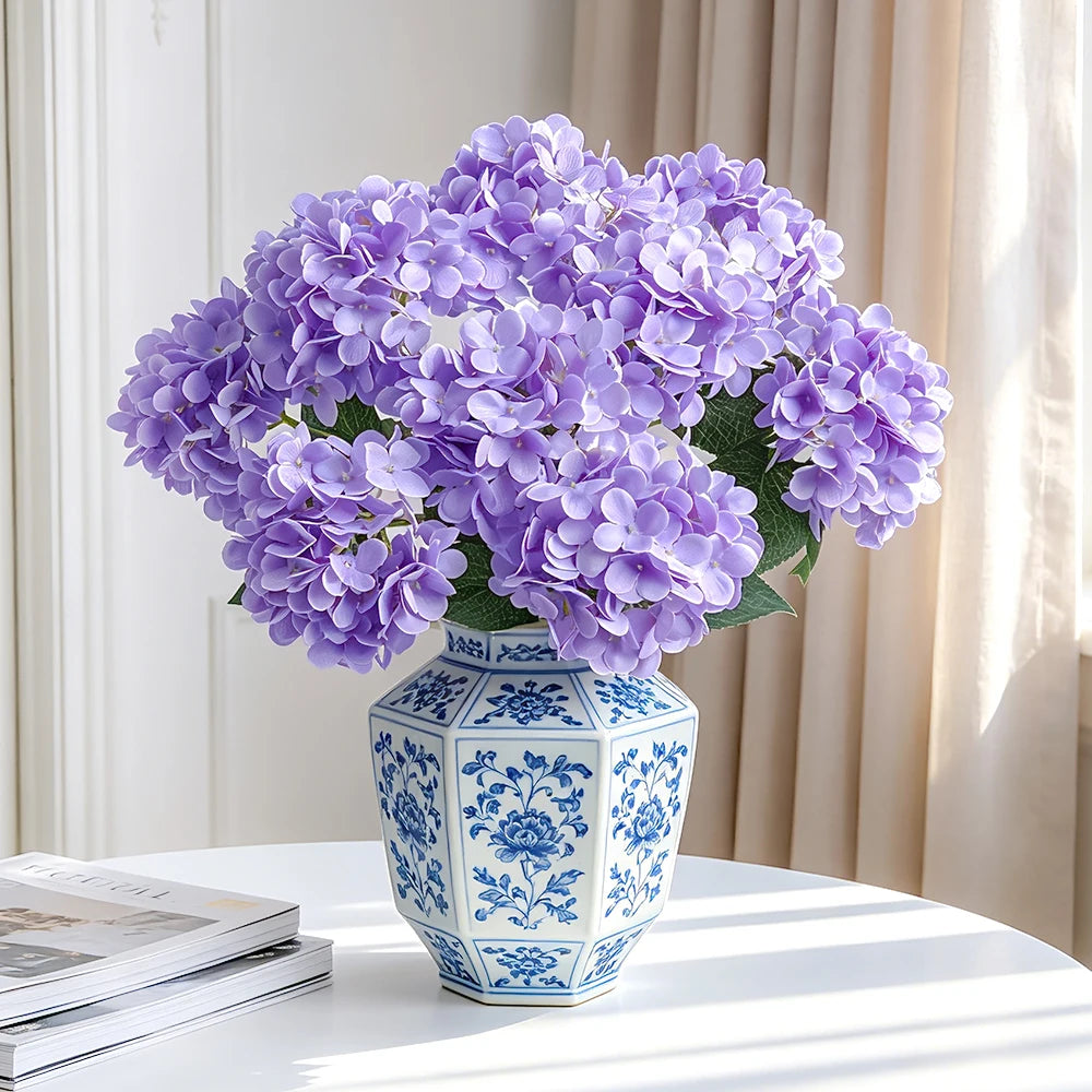 A bouquet of purple artificial hydrangea silk flowers in a blue and white patterned vase on a white table.