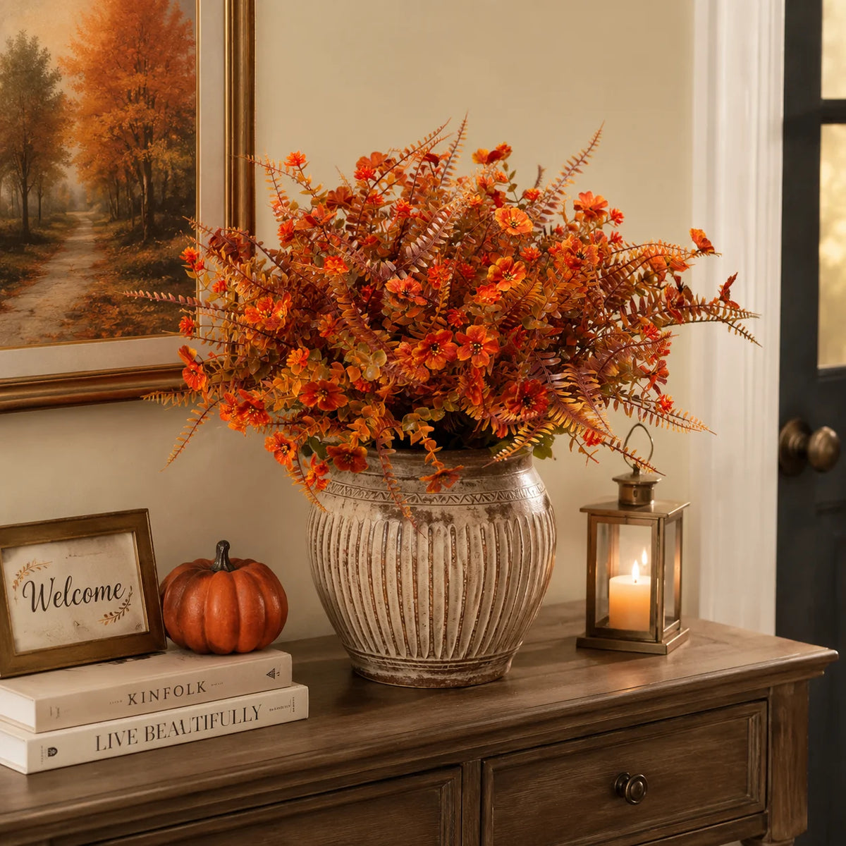 Eine Vase mit künstlichen Herbstblumen in Orange- und Rosttönen steht auf einem Holzschrank neben einer Kürbis- und gerahmten