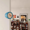 A blue modern resin wall clock is mounted on a white wall next to framed artwork.