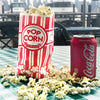 A red and white striped paper bag of popcorn next to loose popcorn on a green checkered table.