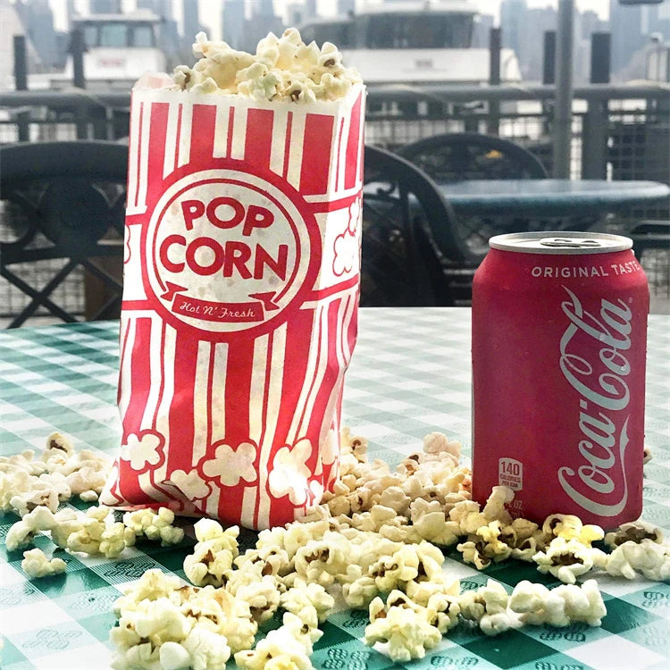 A red and white striped paper bag of popcorn next to loose popcorn on a green checkered table.