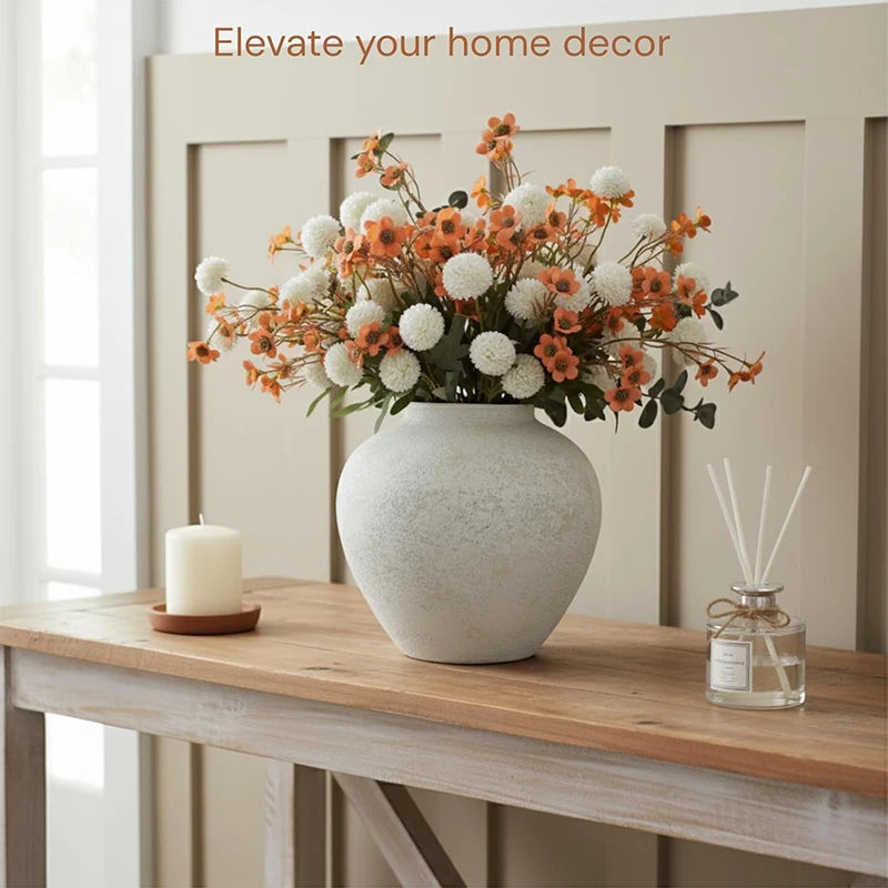 A rustic textured round vase filled with orange and white floral arrangements sits on a wooden table.
