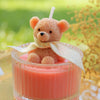 A fluffy orange teddy bear candle in a glass jar with a cream-colored ribbon, set against a green outdoor background.