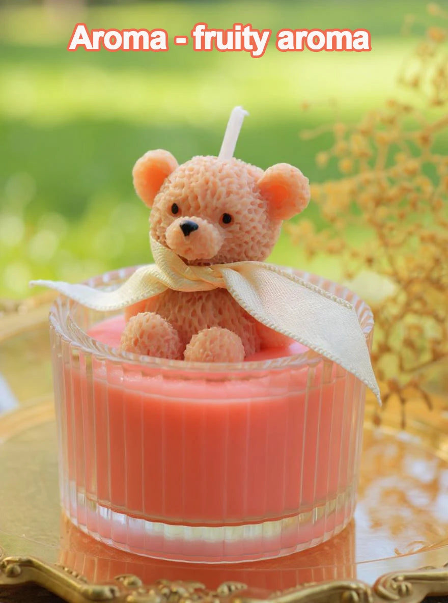 A fluffy orange teddy bear candle in a glass jar with a cream-colored ribbon, set against a green outdoor background.