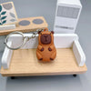 Pendentif de capybara orange sur un porte-clés, exposé sur un plateau en bois clair avec une boîte rectangulaire blanche en a