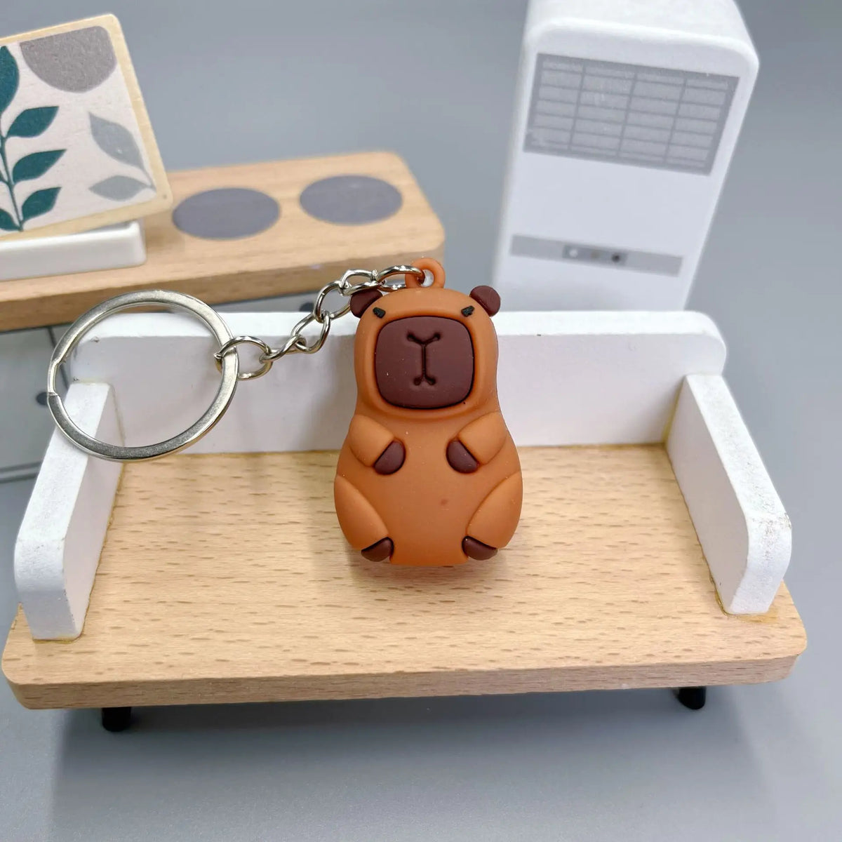 Pendentif de capybara orange sur un porte-clés, exposé sur un plateau en bois clair avec une boîte rectangulaire blanche en a