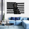 A dark dog is centered on a black and white striped American flag tapestry displayed on a white wall.