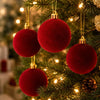 Three deep red velvet flocked Christmas ball ornaments hanging on a green fir tree.