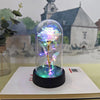 Une rose de verre éternelle avec une lumière LED, présentée sur une base noire, avec un arrière-plan flou d'église et de pays