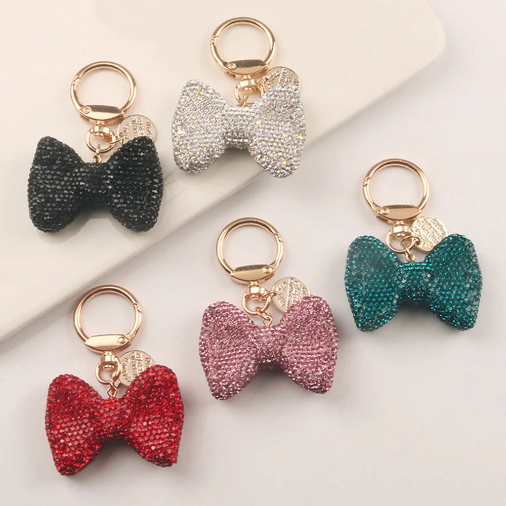 A collection of five sparkling rhinestone bow keychains in black, silver, pink, red, and teal.