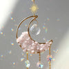 A gold moon-shaped crystal suncatcher featuring pink quartz and iridescent prisms.