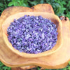Purple tumbled gemstone chips in a wooden bowl resting on a natural wood surface.