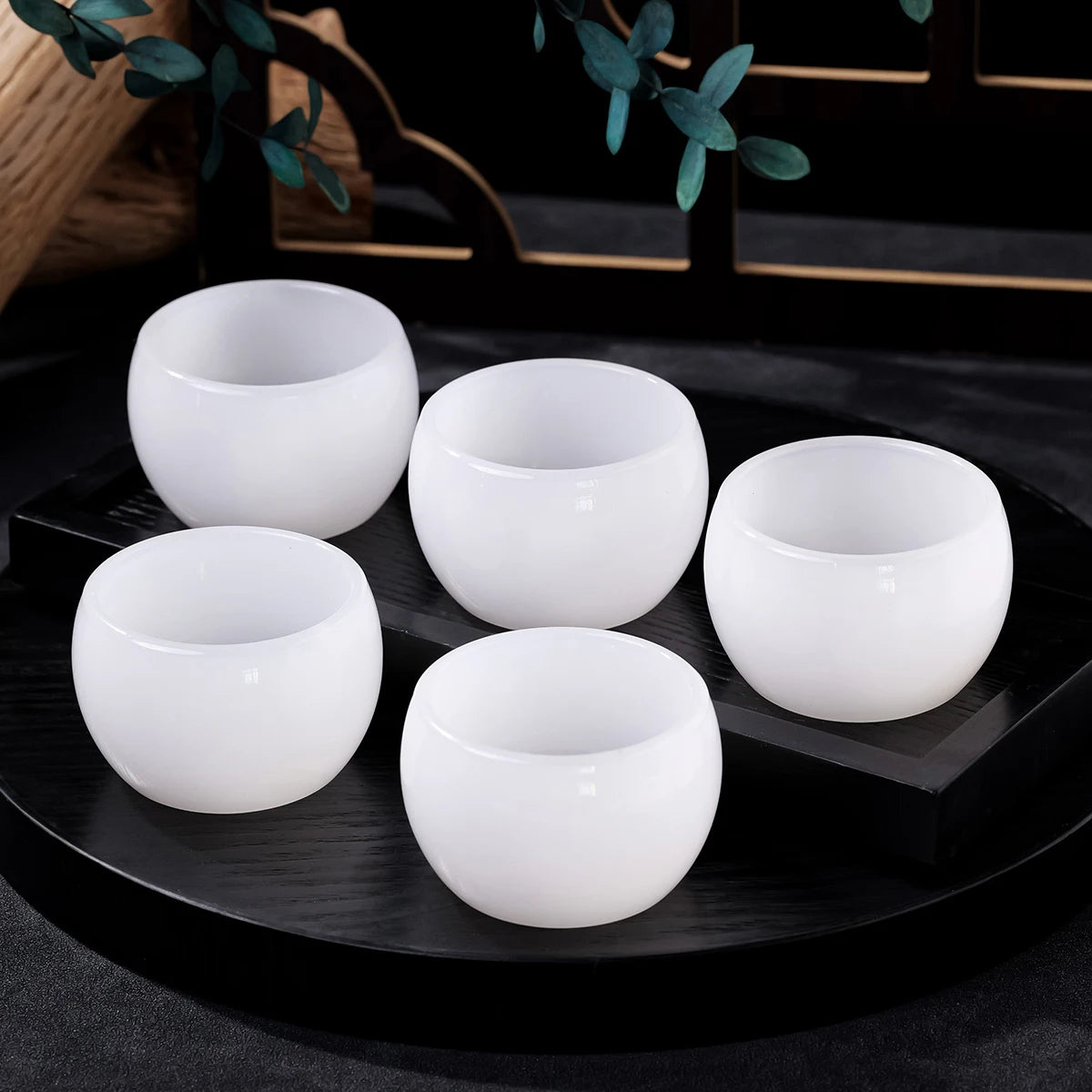 A set of five white ceramic healing bowls displayed on a dark surface with green foliage in the background.