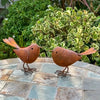 Two rustic metal bird sculptures in brown tones are displayed on a stone surface against a green garden background.