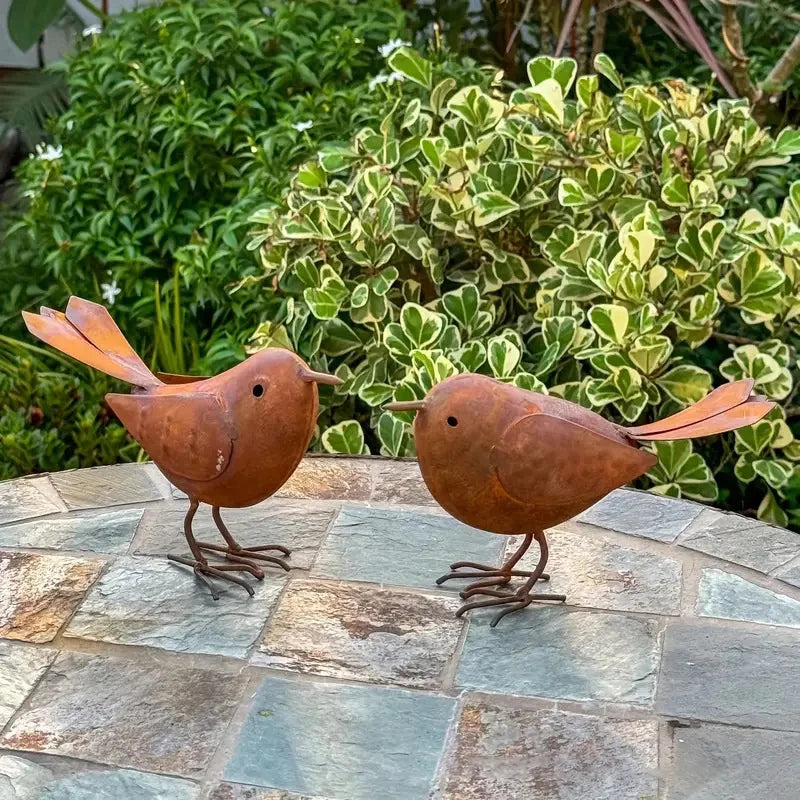 Two rustic metal bird sculptures in brown tones are displayed on a stone surface against a green garden background.