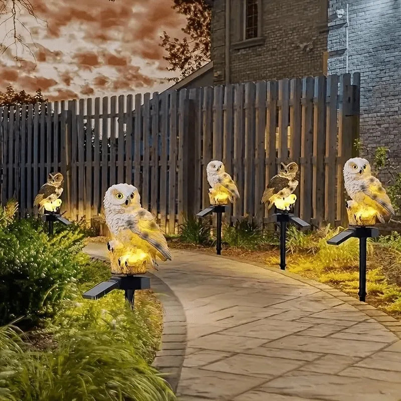 A row of solar-powered owl garden stake lights illuminates owls along a paved pathway in a garden setting.