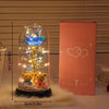 A glass dome containing a glowing golden rose and sparkling lights rests on a fluffy white surface next to its pink packaging
