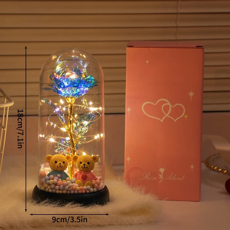 A glass dome containing a glowing golden rose and sparkling lights rests on a fluffy white surface next to its pink packaging