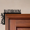 A dark metal plaque with the word BATHROOM is mounted on a light wall above a wooden door frame.