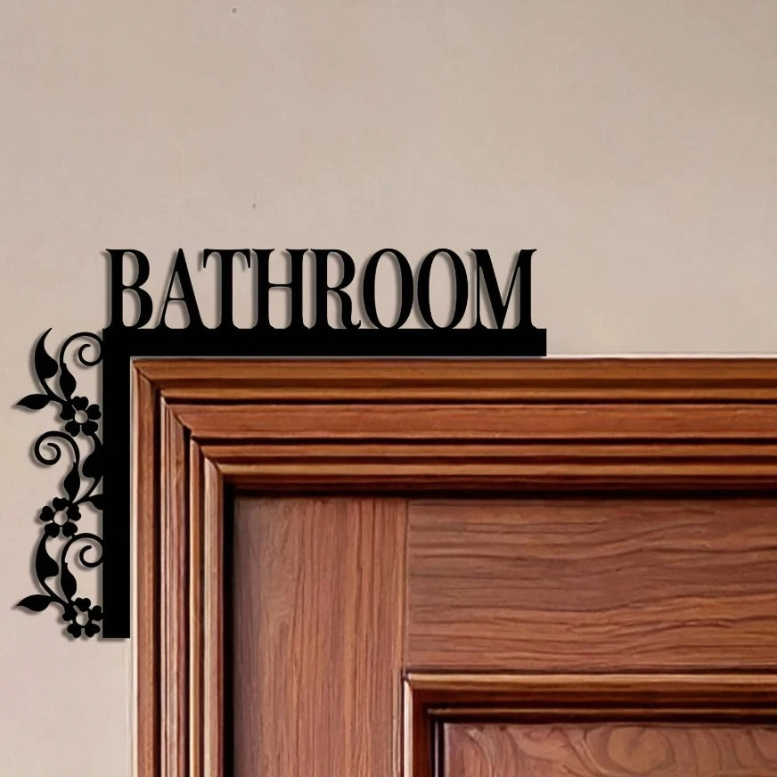 A dark metal plaque with the word BATHROOM is mounted on a light wall above a wooden door frame.