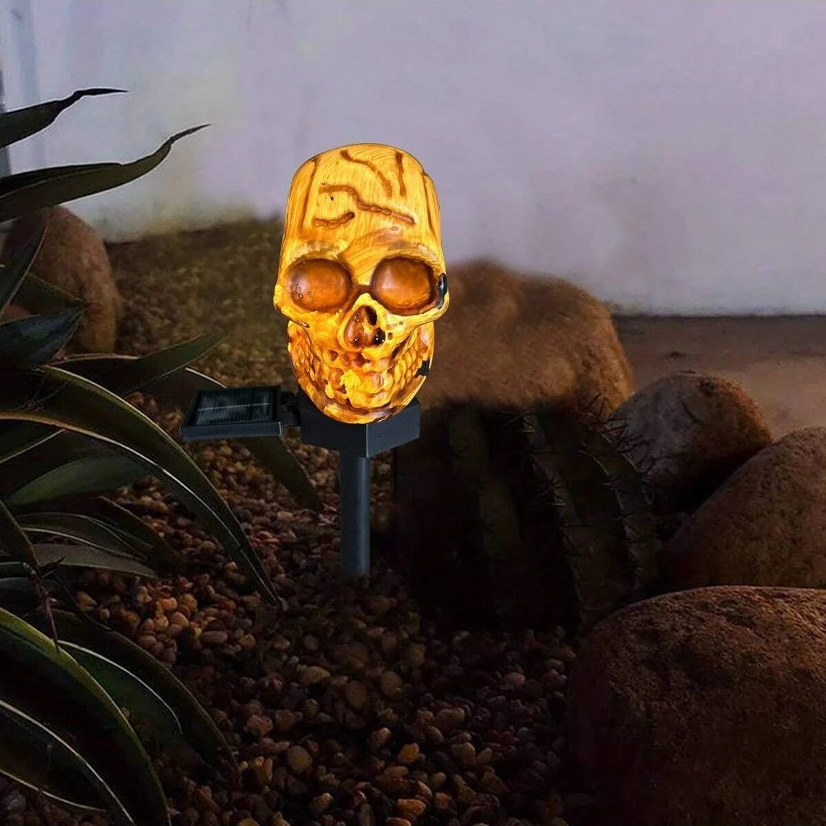 Un luminaire crâne en bois sculpté avec une lueur orange, placé dans un jardin entouré de rochers sombres et de feuillage.