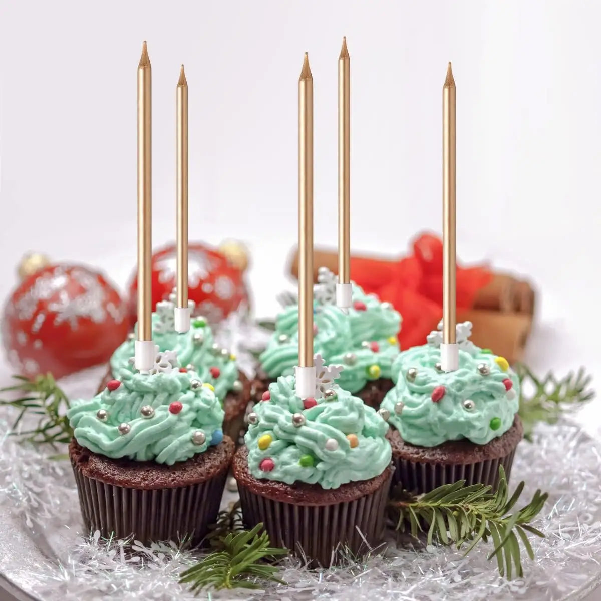 A set of metallic gold birthday cake candles with holders displayed on a white surface surrounded by green foliage.