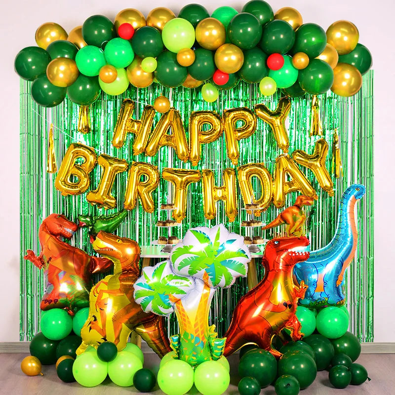 A balloon arch setup featuring foil balloons in green and gold, a tinsel curtain backdrop, and dinosaur-themed props.