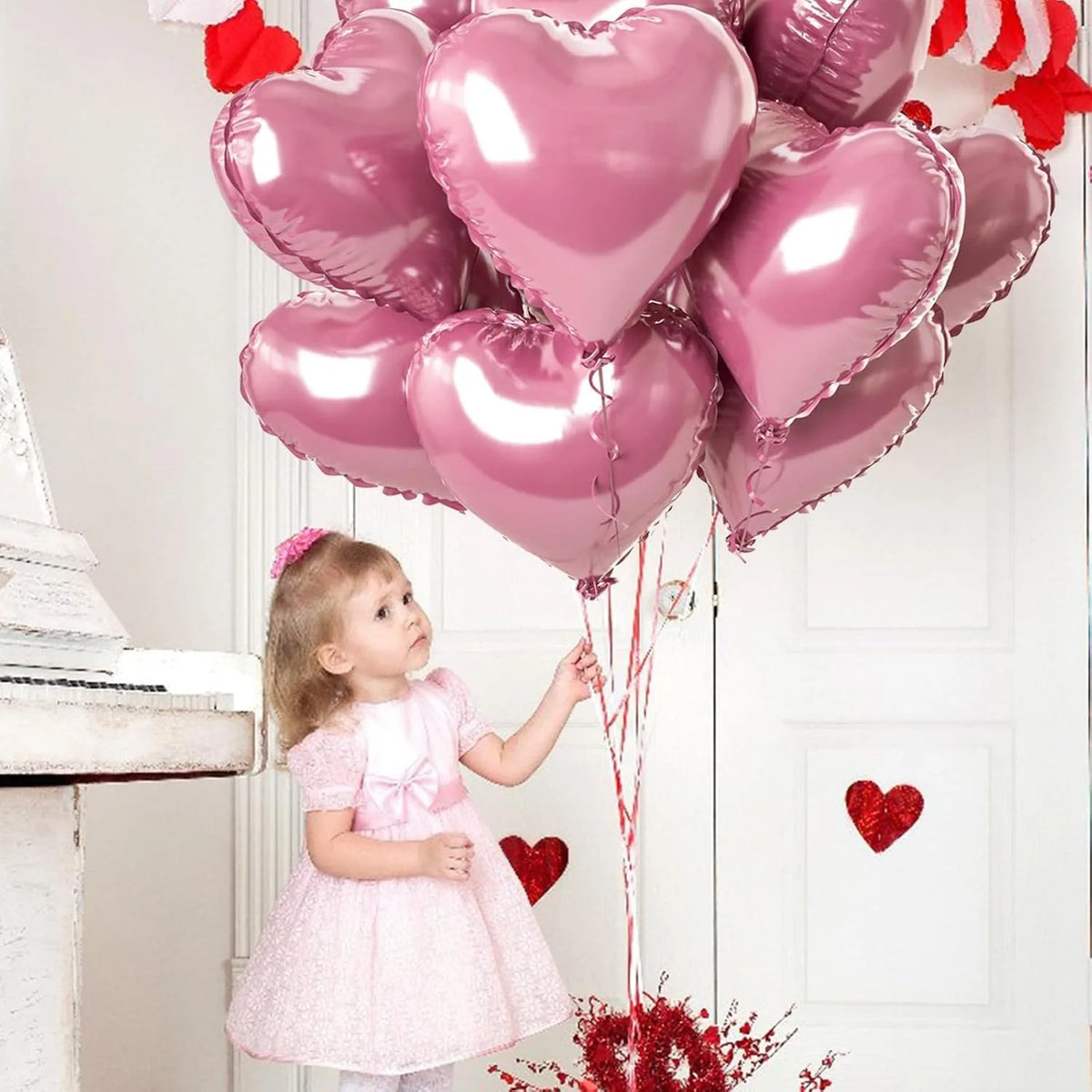 Ein junges Mädchen in einem rosa Kleid blickt auf einen Haufen rosa Herzballons über dem Kamin.