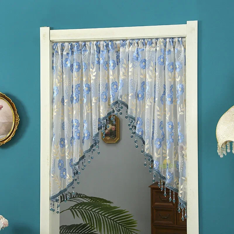 A sheer blue and white embroidered lace door curtain with tassels hanging in a white frame.