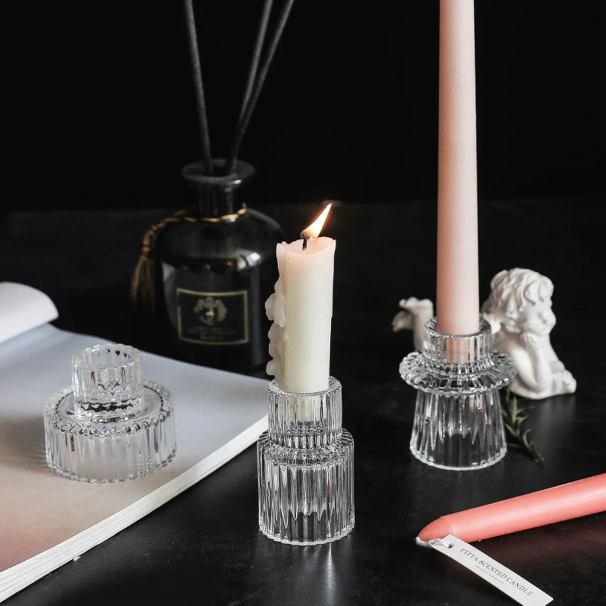 Un portavelas de cristal transparente en forma de pilar azul verdoso junto a una vela rosa y una pequeña estatua blanca.