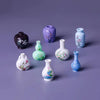 A collection of eight mini resin vases in various shades of purple and blue with floral patterns displayed on a gray surface.