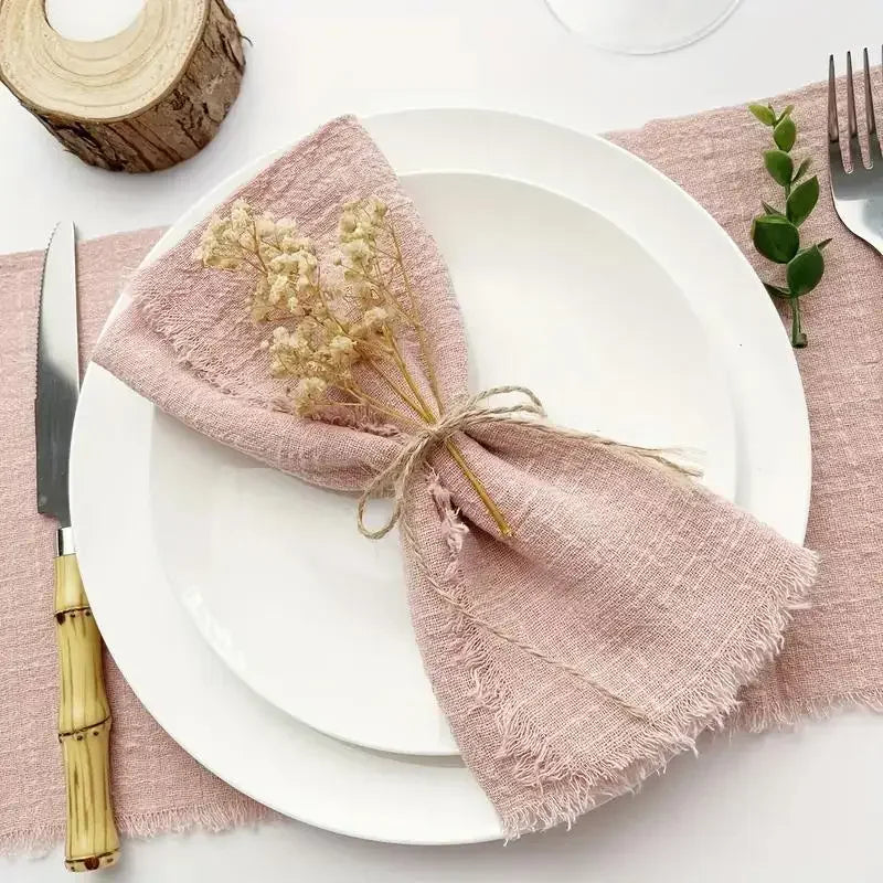 A stack of ten handcrafted fringed cotton napkins in a dusty rose color placed on white plates.