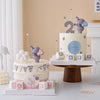 A tiered birthday cake decorated with gray elephant toppers and pastel accents on a white table.