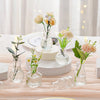 A collection of diamond faceted glass bud vases holding light yellow and peach roses and greenery on a wooden surface.