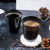 Dunkler Kaffee in einer Tasse auf weißer Untertasse mit Kaffeebohnen und einem Glas mit ganzen Bohnen.