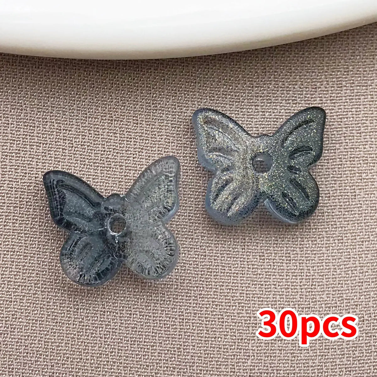 Un juego de tres cuentas de mariposa de vidrio con purpurina gris oscuro mostradas sobre un fondo texturizado.
