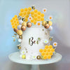 A white cake decorated with yellow honeycomb shapes, gold and silver spherical accents, and small bee figures.