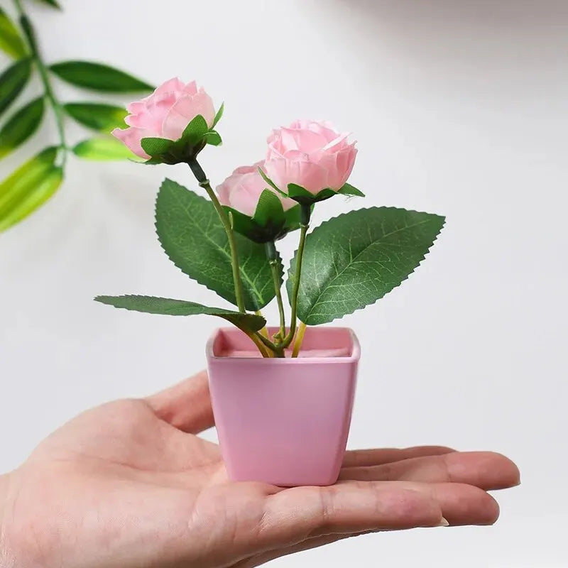 A hand holds a potted artificial pink rose plant with green leaves against a white background.
